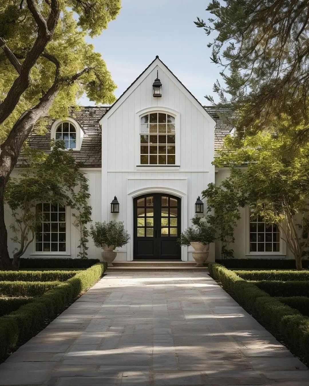 A charming white farmhouse with a welcoming arch-top door and symmetrical windows, nestled among lush trees and manicured greenery.