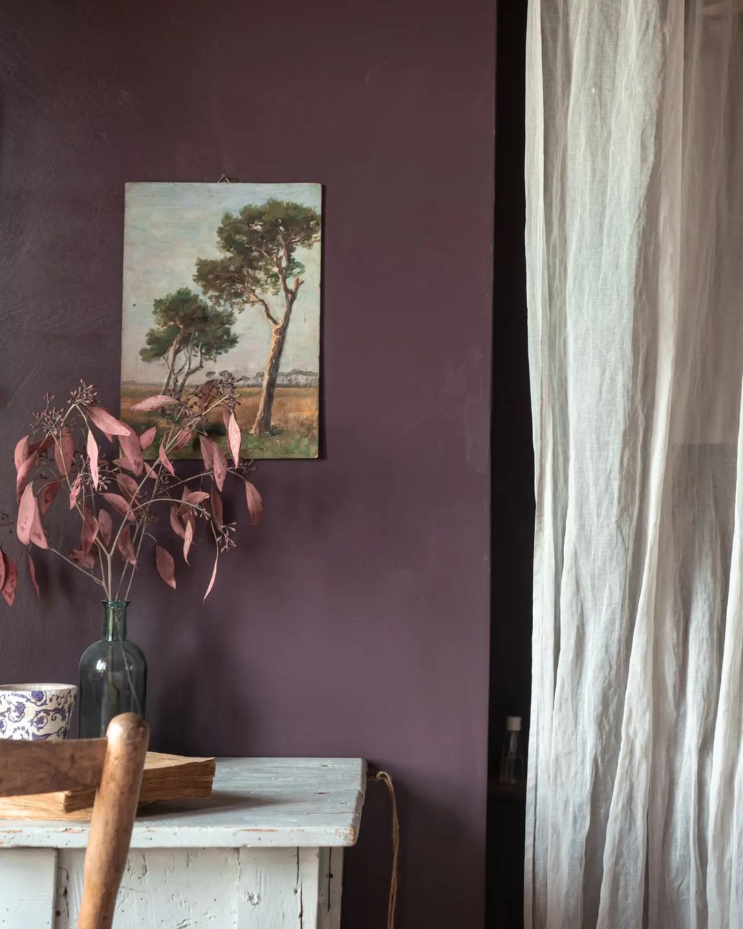 A tastefully composed corner featuring a rustic wooden table with a vase and dried foliage against a deep plum wall, paired with a delicate landscape painting and sheer curtains.