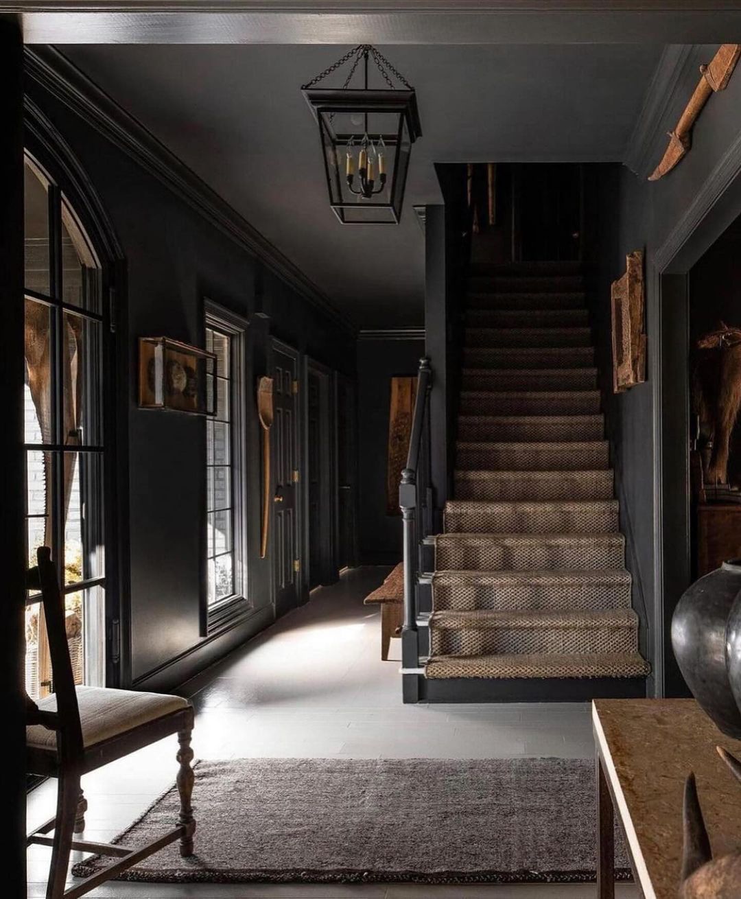 An ambiance of mystery and sophistication envelops the hallway, characterized by its **dark grey palette**, where various old-timey artifacts adorn the walls and an *antique-style lantern* hangs overhead, casting a soft glow over the carpet-lined staircase.