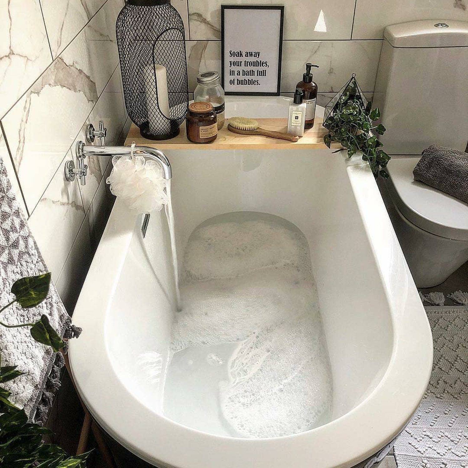 A modern bathroom featuring a freestanding bathtub with foamy water, surrounded by plants and bath accessories