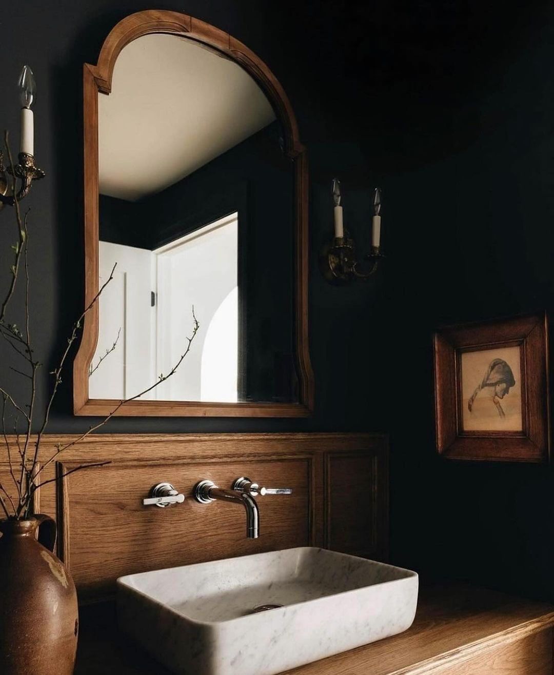 A sophisticated dark-toned bathroom featuring a wooden vanity with a marble sink, wall-mounted taps, and a vintage mirror framed in wood