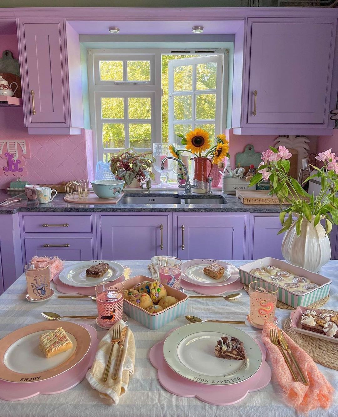 A cozy and whimsical kitchen with pastel purple cabinets, a playful pink tile backsplash, and a sunlit window.