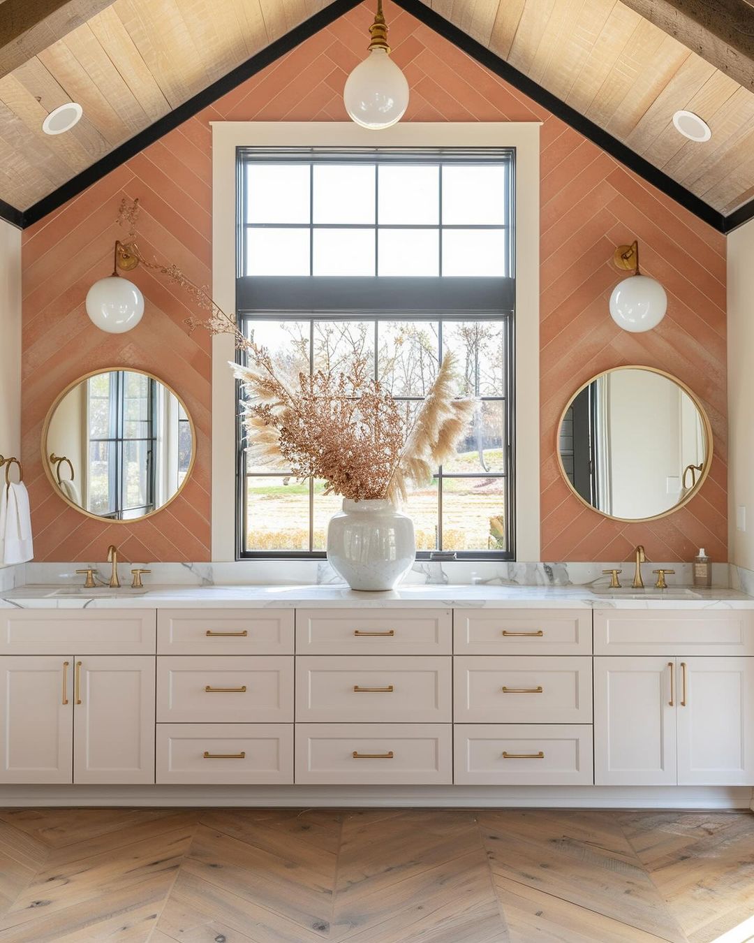 A chic and sleek bathroom featuring herringbone parquet flooring, a terracotta tiled accent wall, dual round mirrors, and a modern white vanity with gold hardware.