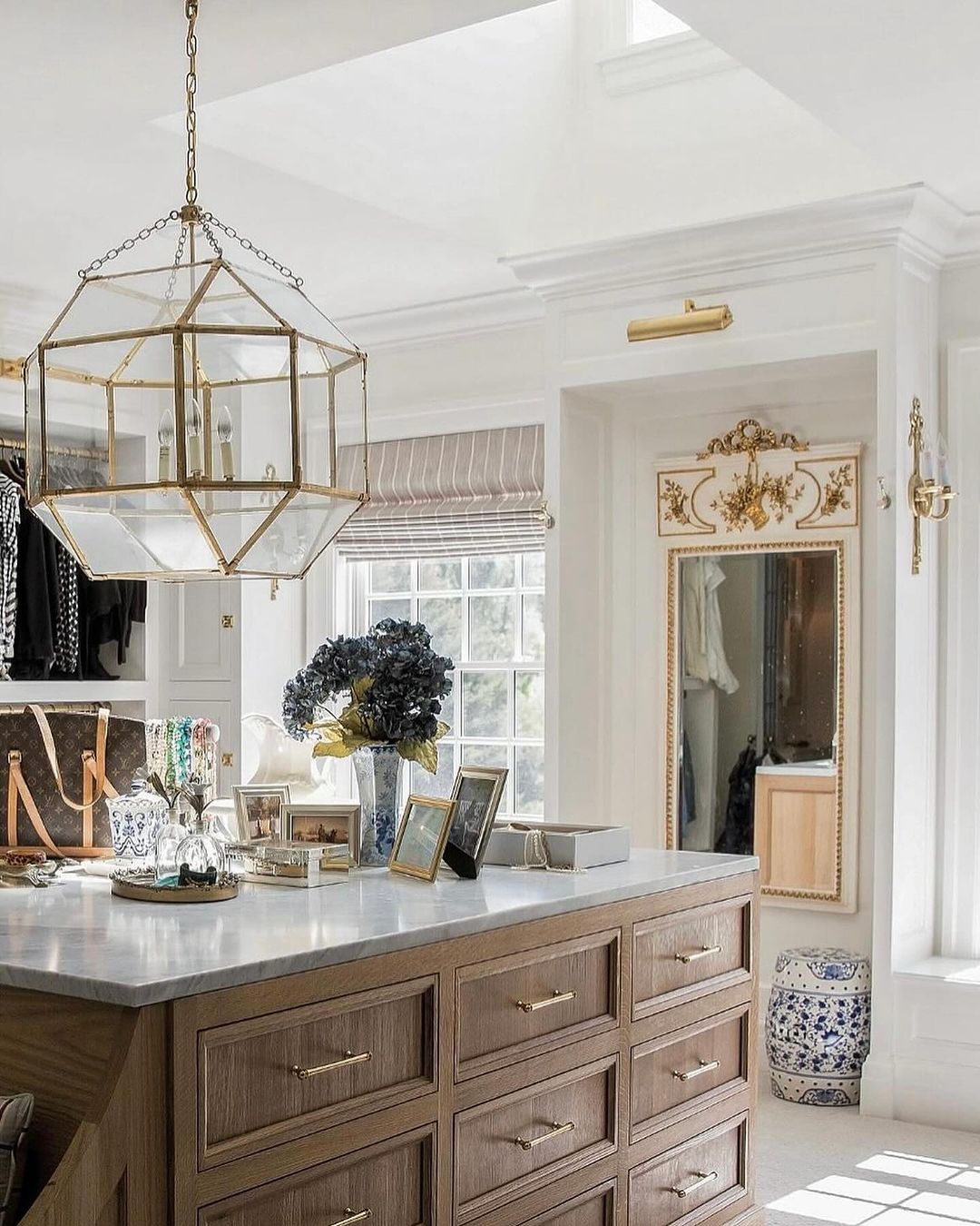 A chic and well-organized closet space featuring a wooden island with brass handles, marble countertop, and a pendant light