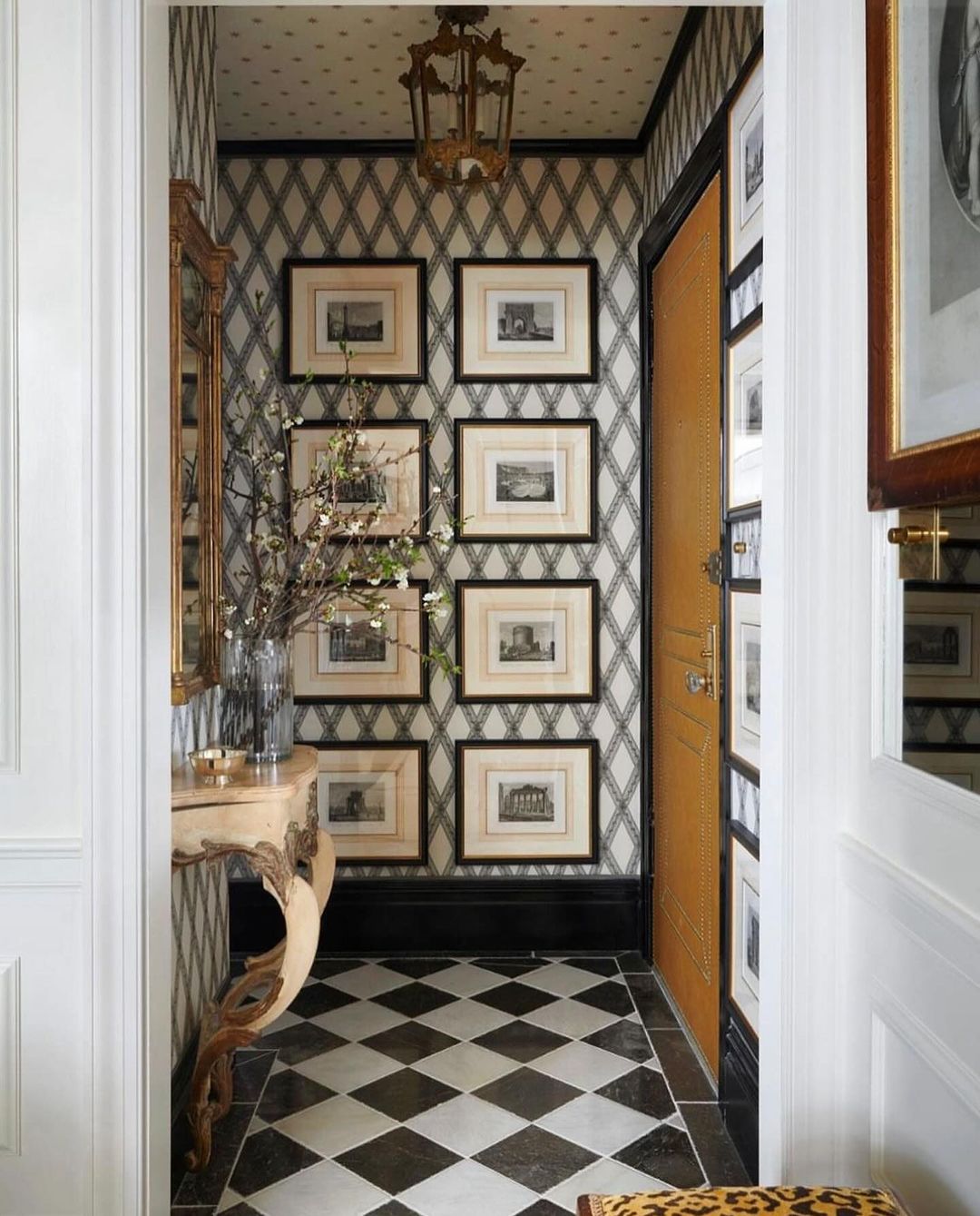 A classic meets eclectic foyer featuring a bold harlequin floor and diamond-patterned wallpaper, complemented by a curvaceous wooden console and a cluster of traditional framed artwork.