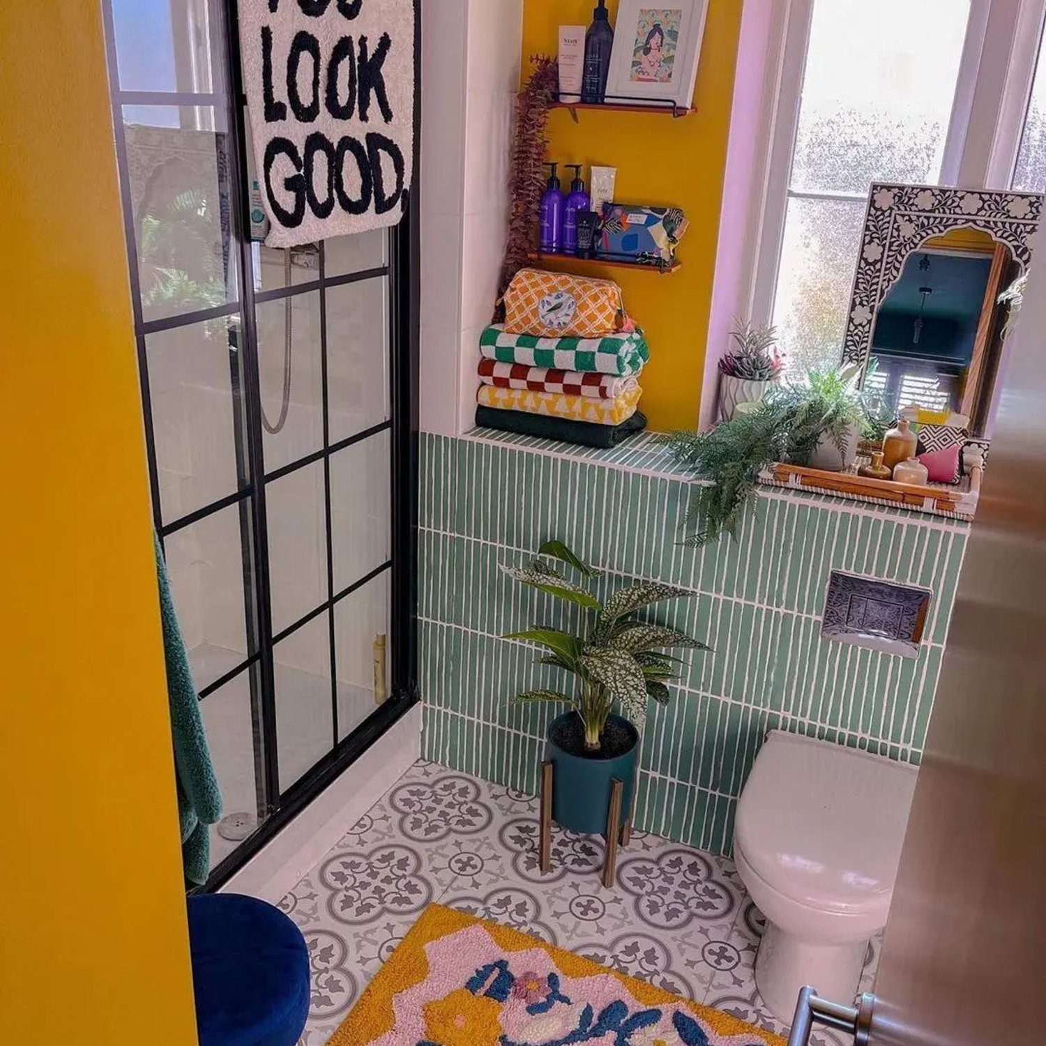 A vibrant and eclectic bathroom featuring a patterned yellow floor, teal striped walls, a clear glass shower partition, and playful accents such as a 'YOU LOOK GOOD' towel and a colorful rug.