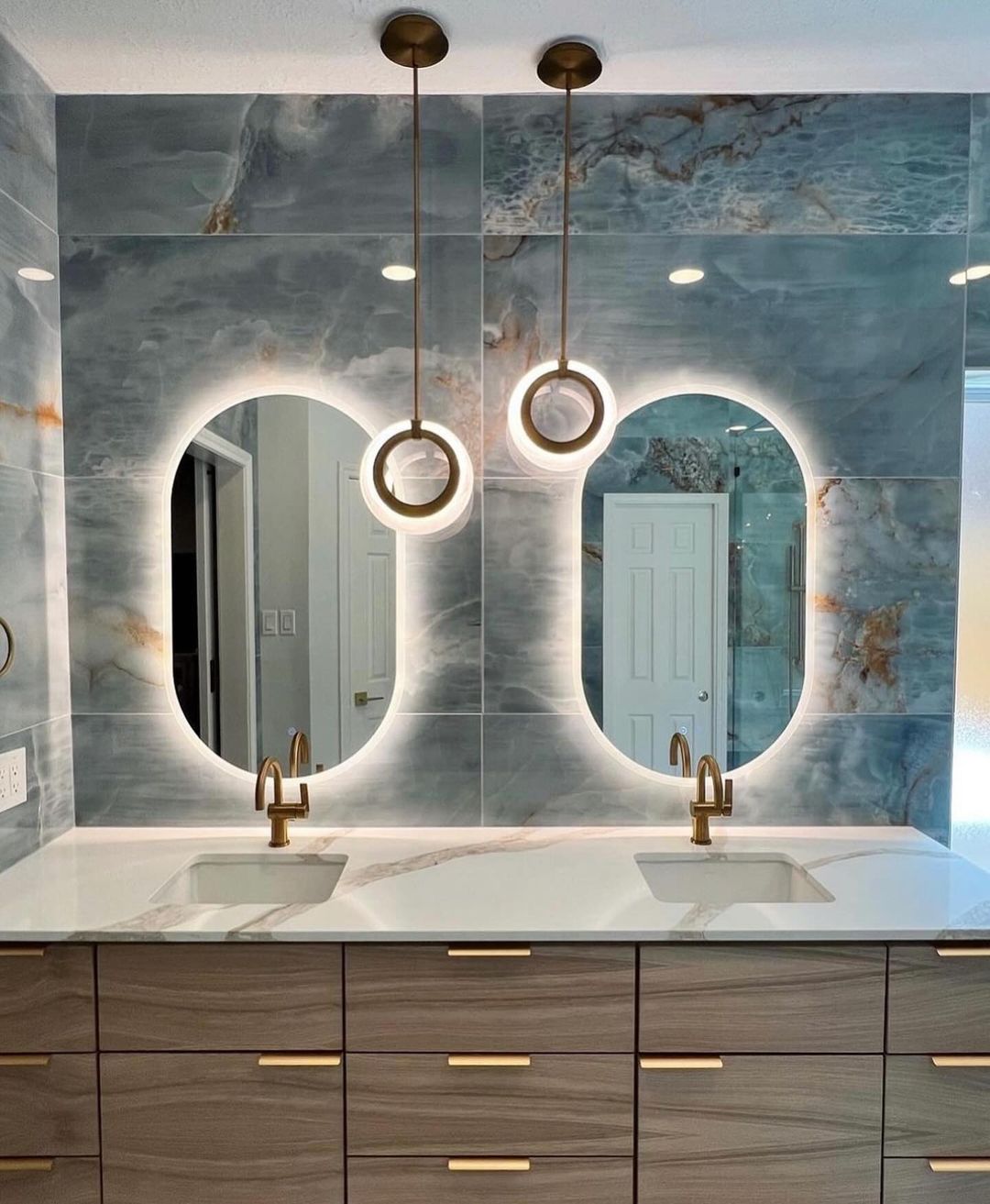 A contemporary bathroom featuring dual arched mirrors set against a marbled wall, with circular pendant lights