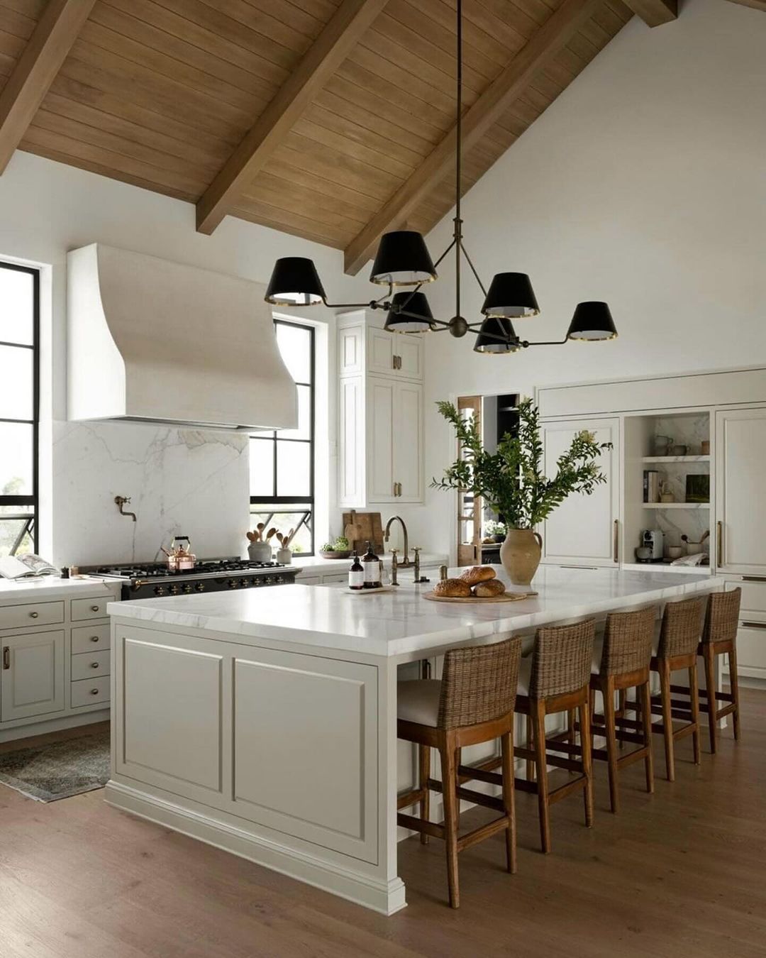 A sleek modern kitchen featuring exposed wooden beams, contrasting black pendant lights, and a central island with woven bar stools