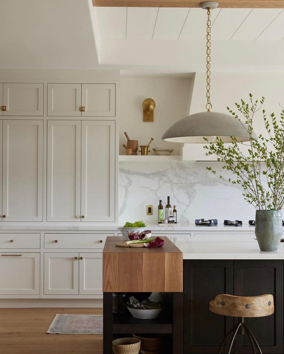 A modern and warm kitchen space featuring white cabinetry, marble backsplash, and a contrasting dark island with a unique saddle stool