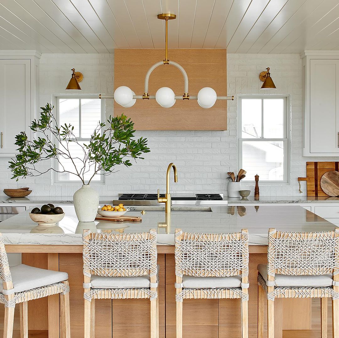 A modern kitchen featuring exposed wooden beams, woven bar stools, and brass fixtures