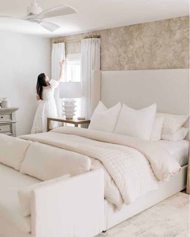 A person adjusting the curtains in a well-organized bedroom with a neutral color palette