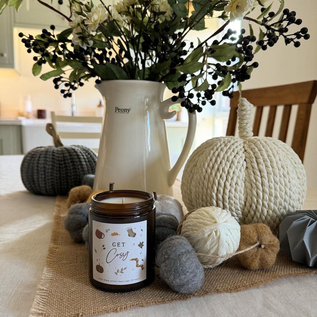 A cozy autumn centerpiece arrangement featuring a cream pitcher with flowers, textured pumpkins, and a 'Get Cozy' candle