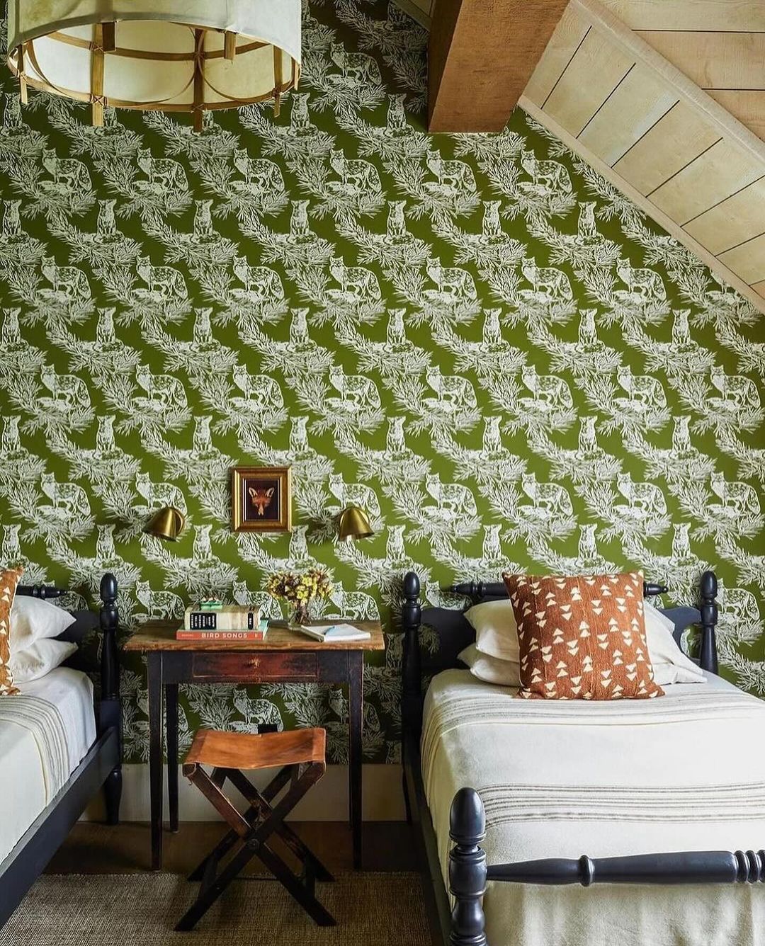 A cozy bedroom featuring a striking green wallpaper with a white botanical pattern, black vintage-style twin beds, and a small rustic desk