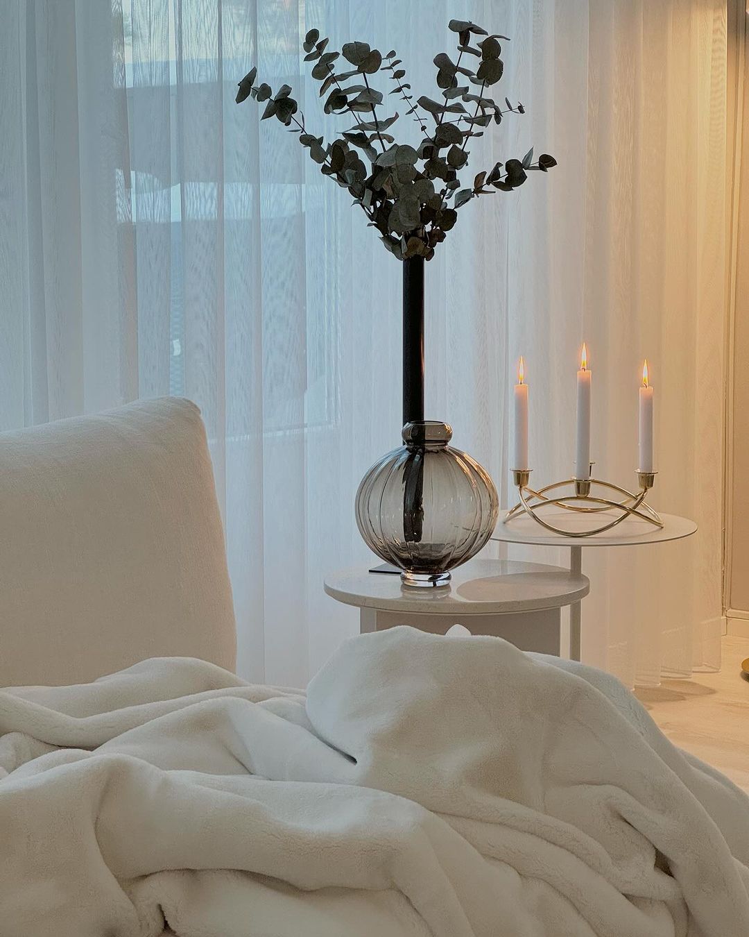 A tranquil nook featuring a plush white blanket, an eucalyptus bouquet in a ribbed glass vase on a side table, and a simple gold candle holder with lit candles.