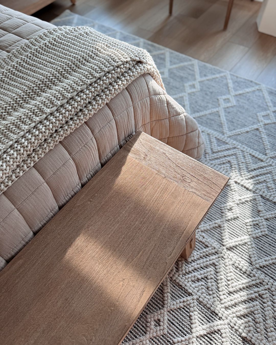 A cozy home corner featuring a plush beige throw over a quilted ottoman, beside a wooden arm of a couch, with a geometric-patterned rug underneath bathed in natural sunlight