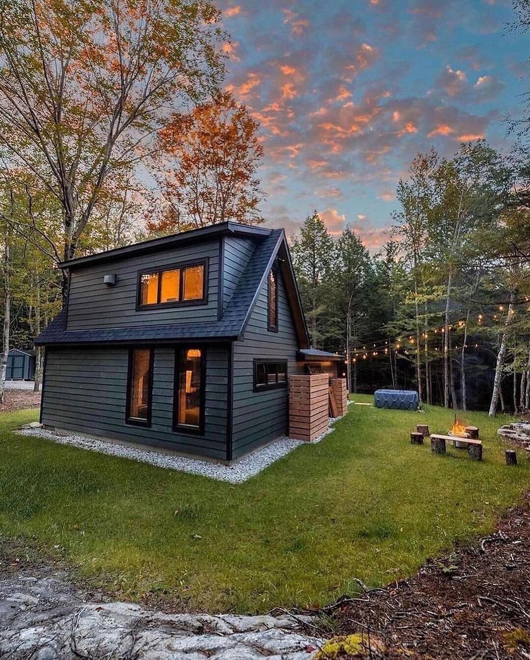 A cozy modern cabin nestled in the woods with warm interior lights and a fire pit outside