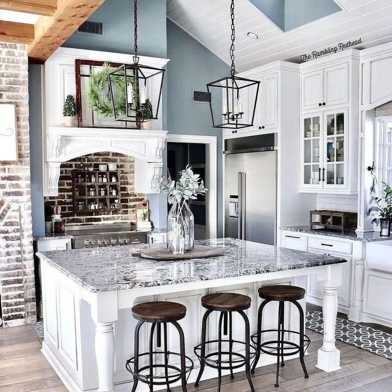 Elegant and spacious kitchen featuring exposed wooden beams, a large center island with granite countertop, modern metal pendant lighting, and a mix of white cabinetry with brick accent walls.