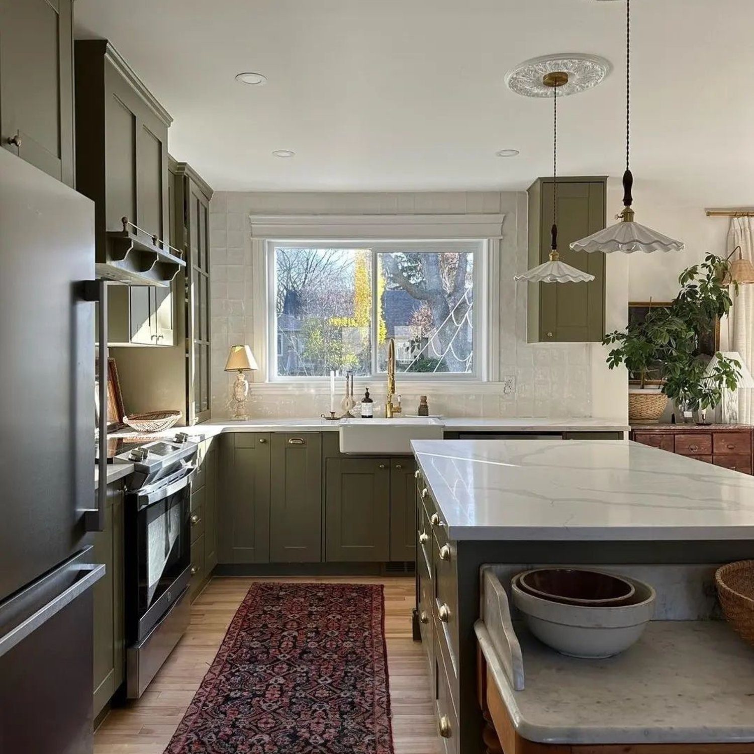 A cozy and elegant kitchen with sunlight filtering through the window, highlighting the olive green cabinets and the warm wooden floors.