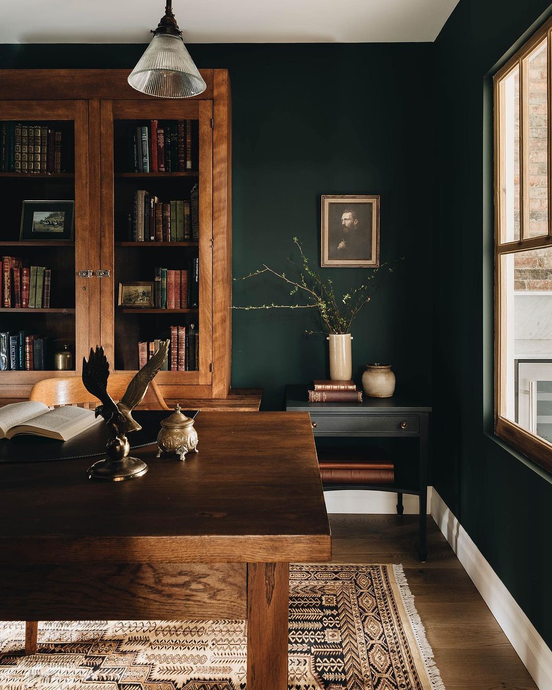 A cozy and inviting home office with dark green walls, a warm wooden desk, and bookshelf, accented by a patterned rug.