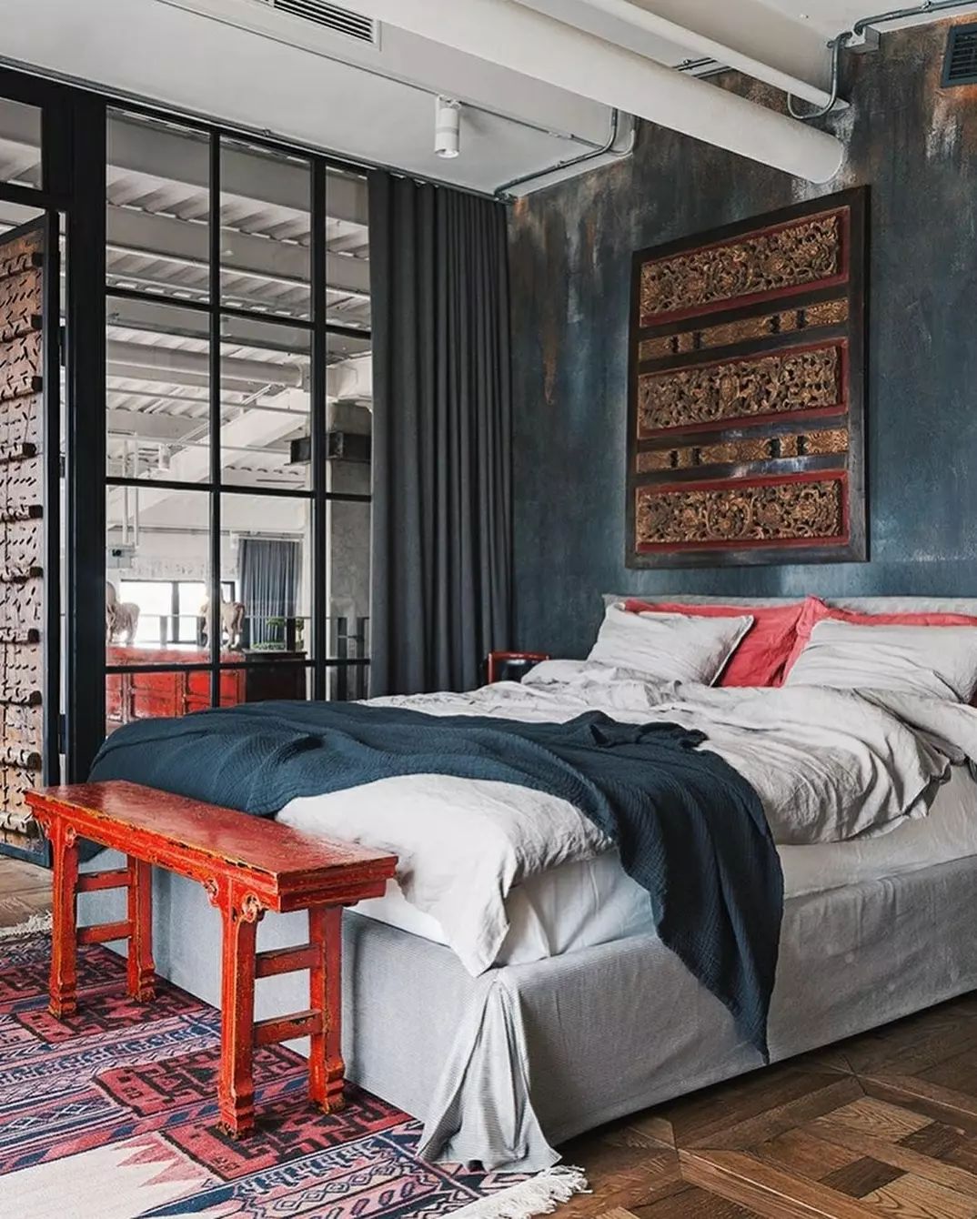 A chic industrial-style bedroom featuring a textured dark wall, a vibrant red bench at the foot of the bed, and a patterned rug