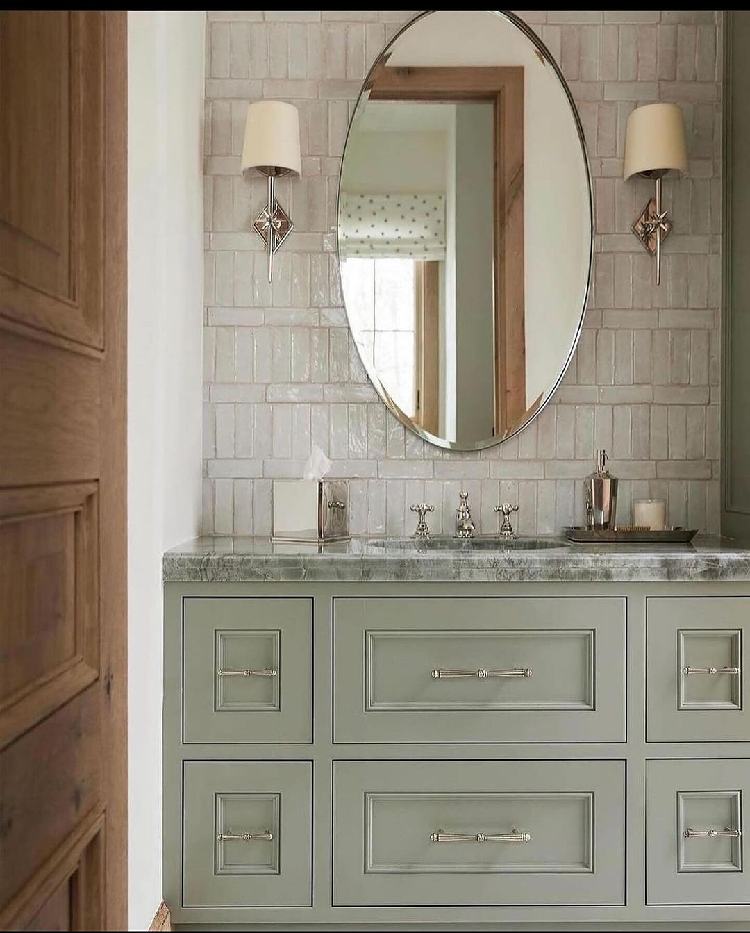 A tastefully designed bathroom with a textured grey tile backsplash, oval mirror, and stylish sconces