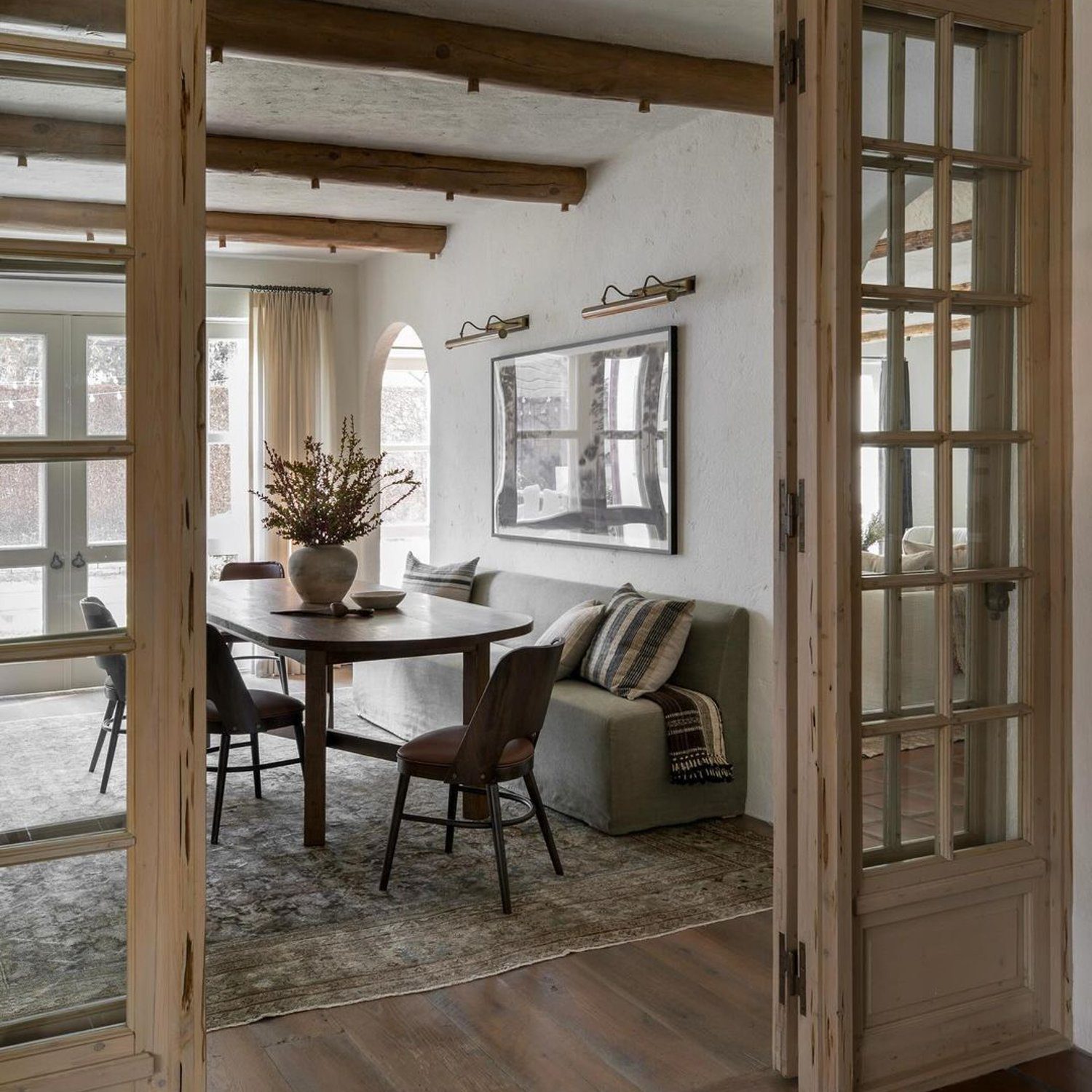 A cozy dining and living area with rustic charm featuring exposed wood beams, white plaster walls, and traditional glass-paned doors.