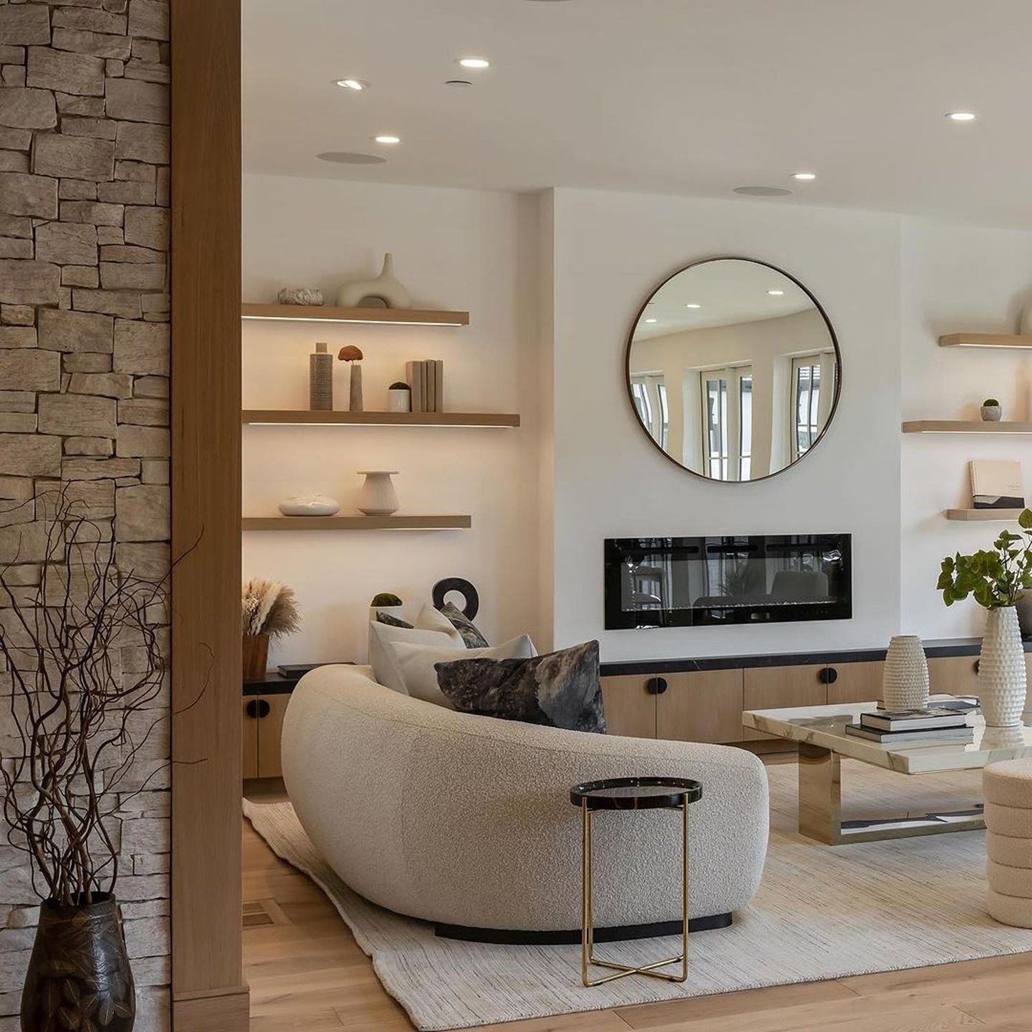 A serene living room design boasting an inviting curved bouclé sofa, accompanied by a modern round side table, and a sleek media unit nestled beneath a clean-lined display of decorative shelving and a round mirror; all tied together with a warm palette of neutral tones, natural wood, and stone accents.