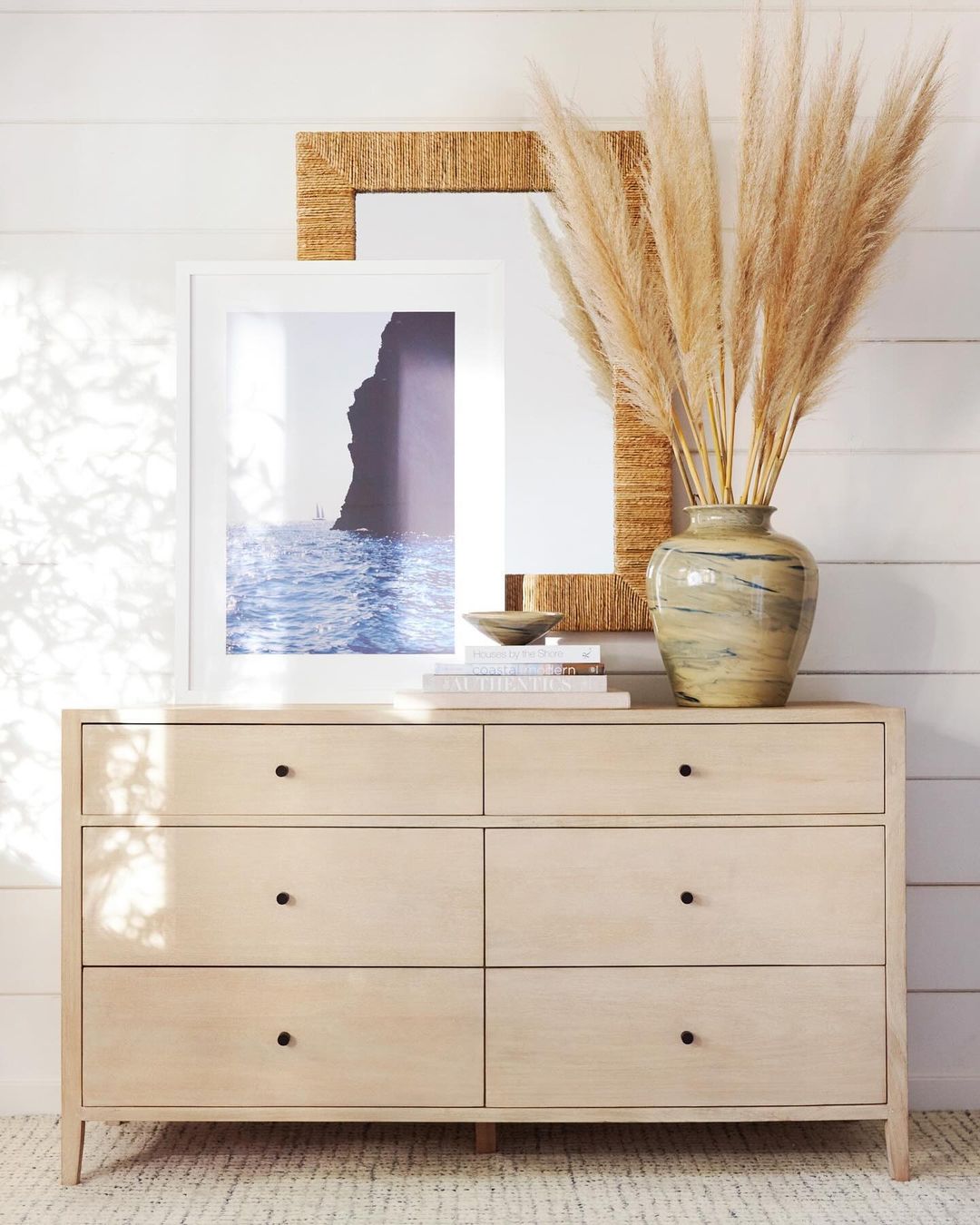 A thoughtfully composed home interior featuring a light wood dresser with six drawers, against a white wall with a textured woven frame artwork and a decorative vase with pampas grass