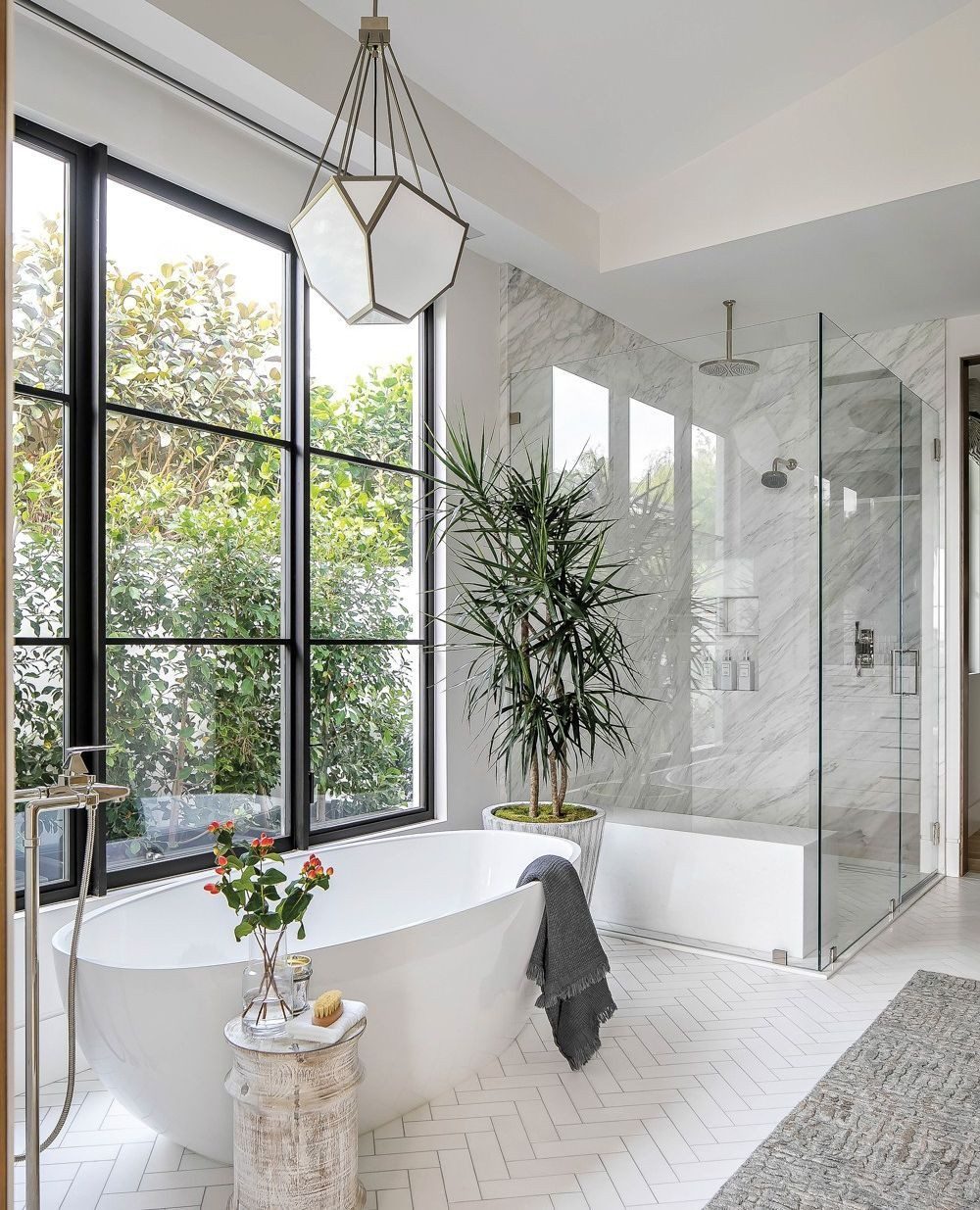 A serene bathroom featuring a free-standing white bathtub near floor-to-ceiling windows with a view of lush greenery, contrasting with the sleek lines of a glass shower and geometric pendant light.