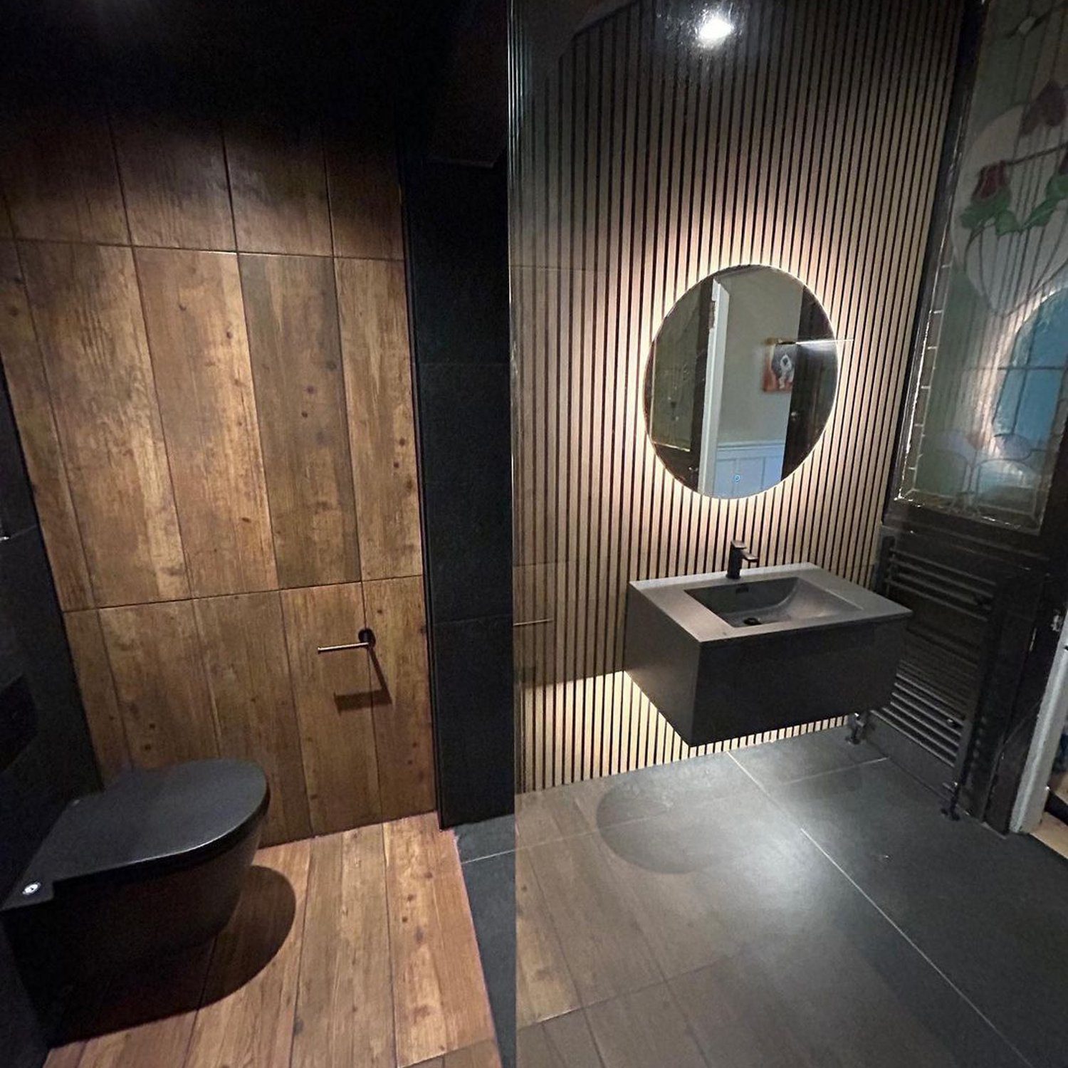 A chic and modern restroom featuring rustic wooden wall panels, a sleek black toilet, and a contemporary basin with a round illuminated mirror.