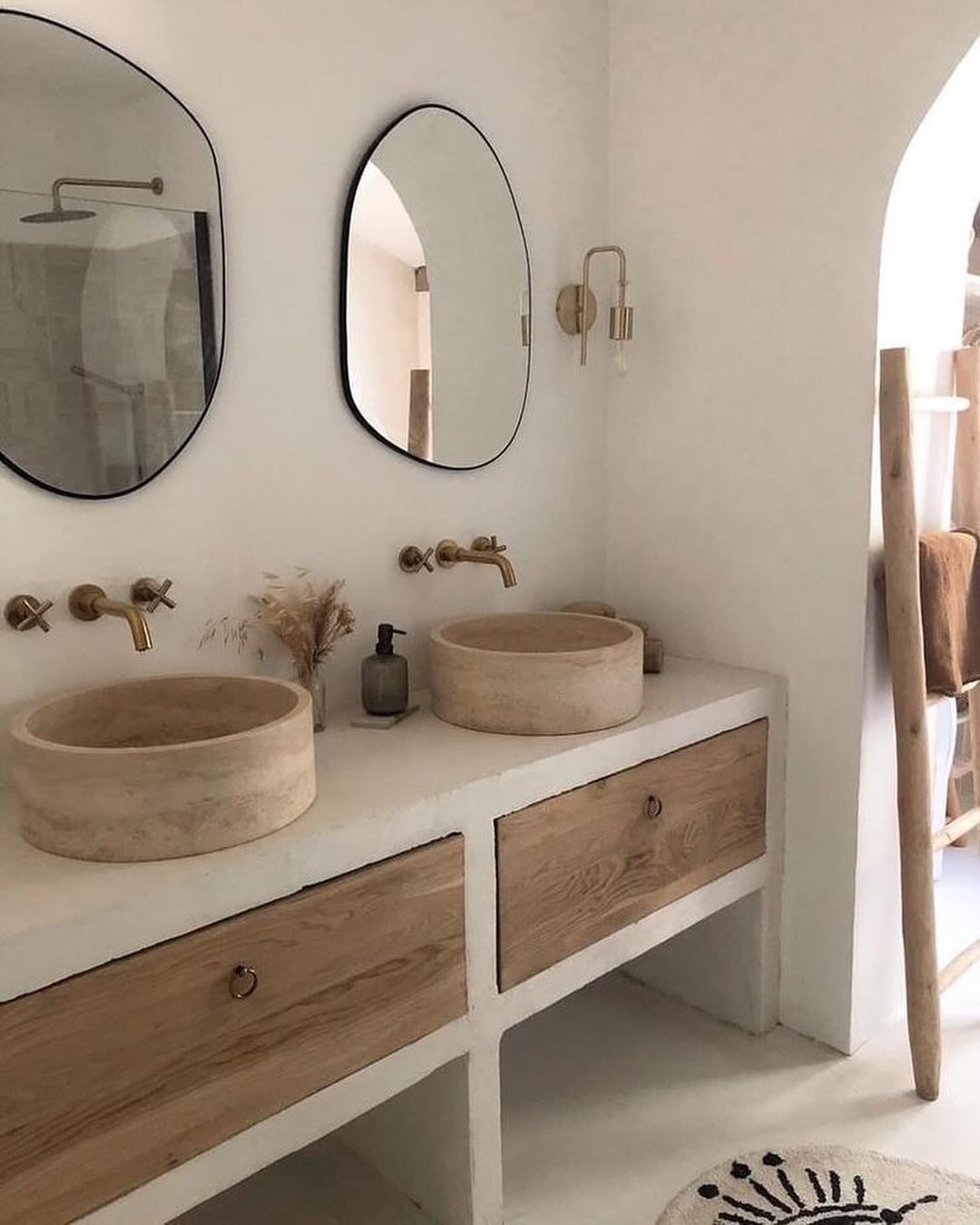 A serene bathroom setting featuring natural tones, with dual stone basins atop a wooden vanity, brass wall-mounted faucets, and oval mirrors.