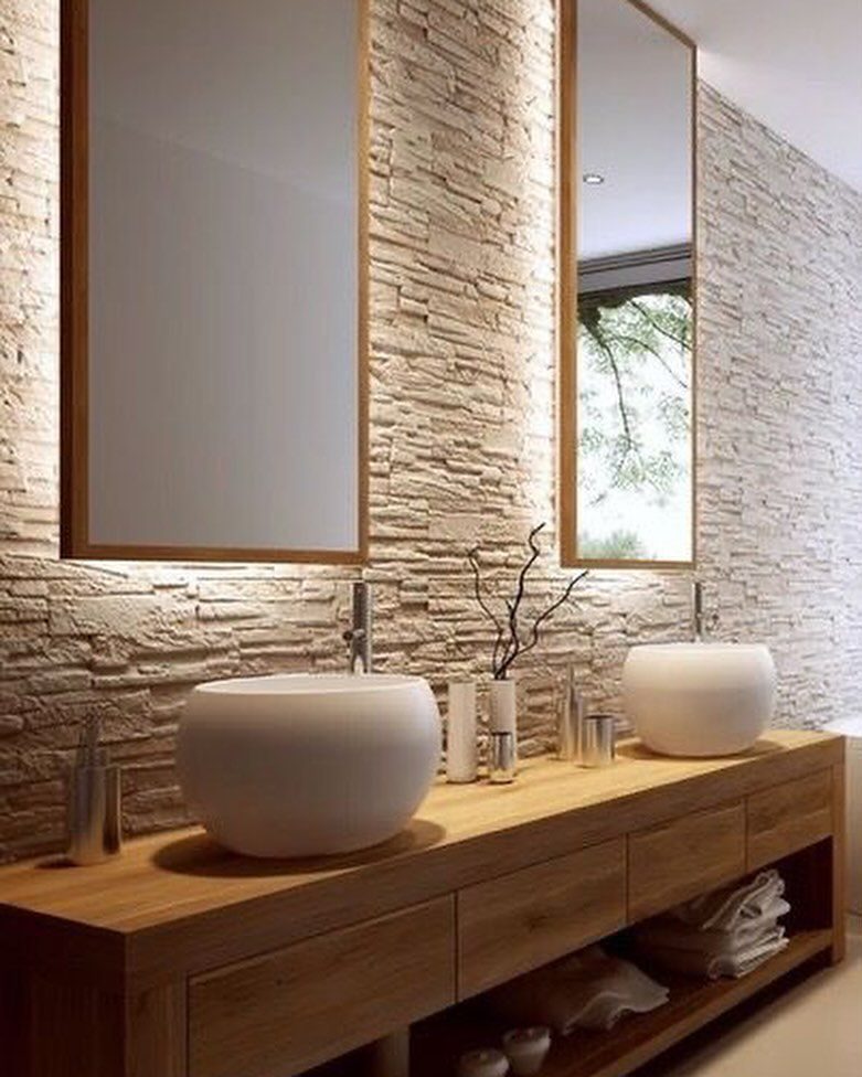 A modern bathroom featuring a textured stone wall, wooden vanity with vessel sinks, and large mirrors reflecting natural light