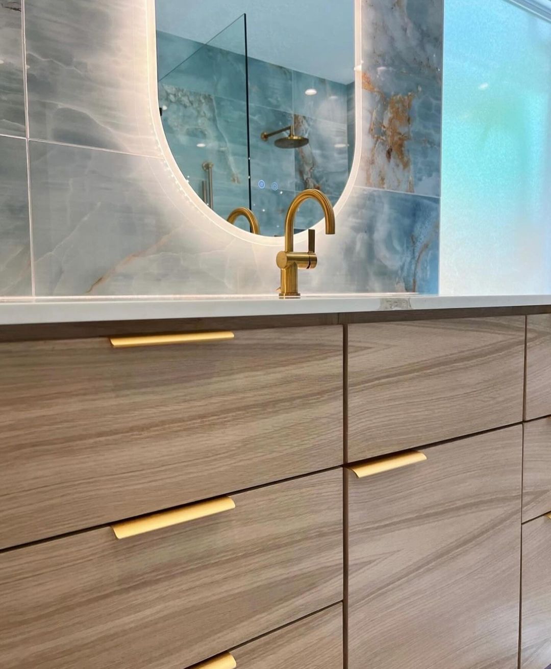 A modern bathroom vanity featuring gold fixtures, wooden cabinetry, and a unique mirror design