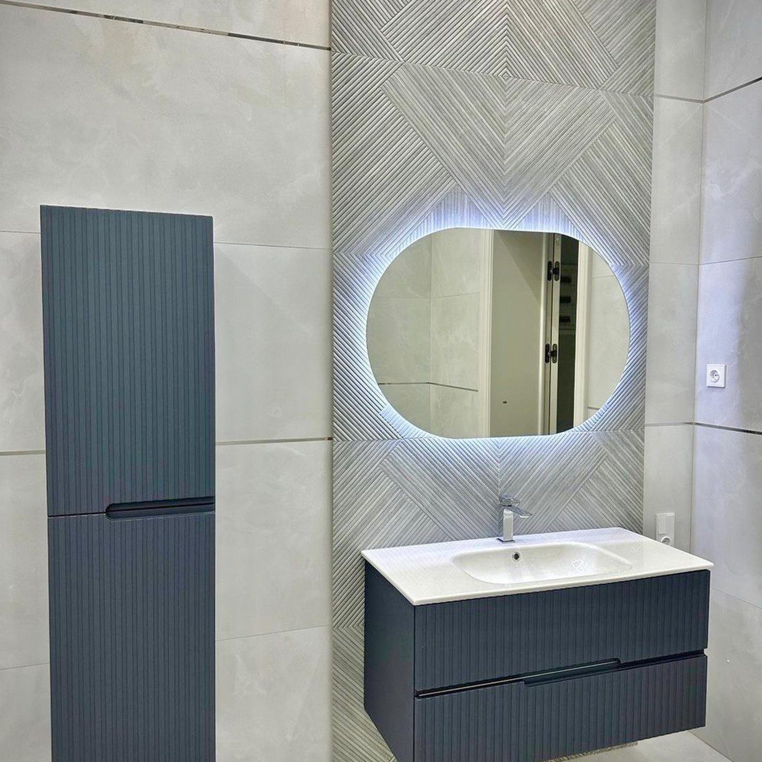 A modern bathroom design featuring a circular illuminated mirror set against a dynamic herringbone tile accent wall, paired with a sleek grey vanity and a complementary tall storage cabinet.