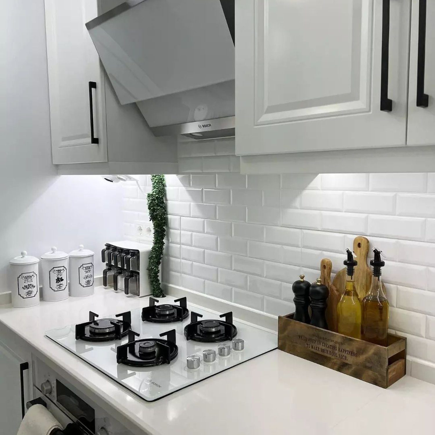 A modern, clean kitchen space with white cabinetry and a subway tile backsplash, featuring a stainless steel gas stove, and stylish kitchen accessories like spice jars, a plant, and decorative oil bottles.