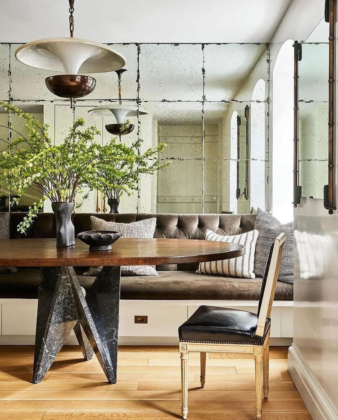 A chic, modern dining room featuring a large antiqued mirror wall reflecting a bold marble table and a classic upholstered chair