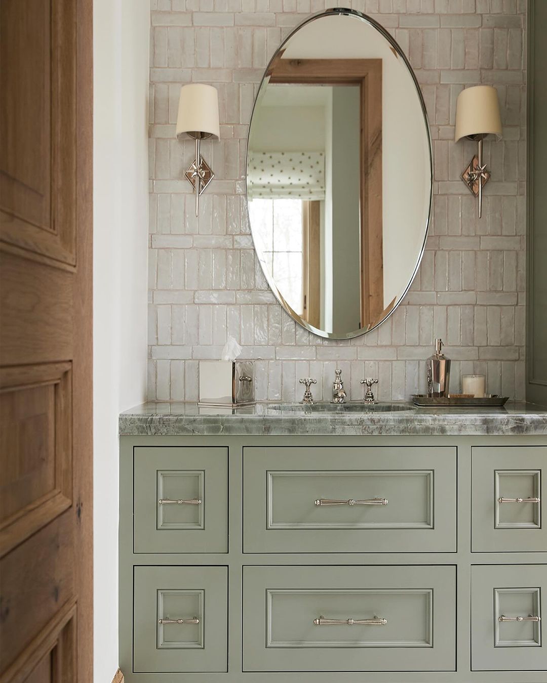 Chic greenish-gray vanity featuring a marble countertop, set against glossy subway tiles, with a classic oval mirror and warm sconce lighting