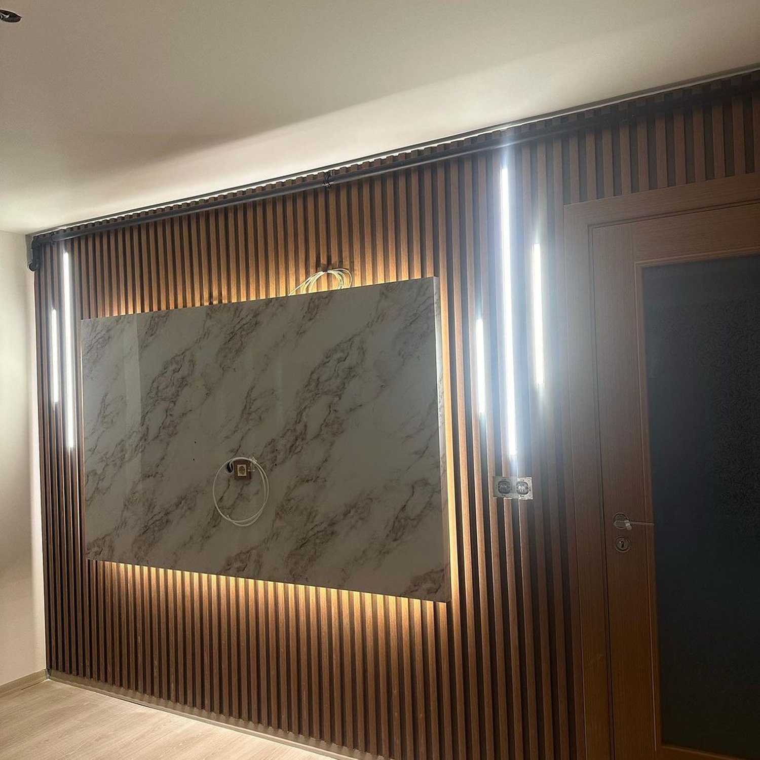 A modern interior wall design featuring vertical wooden slats, a marble inset, and sleek vertical lighting