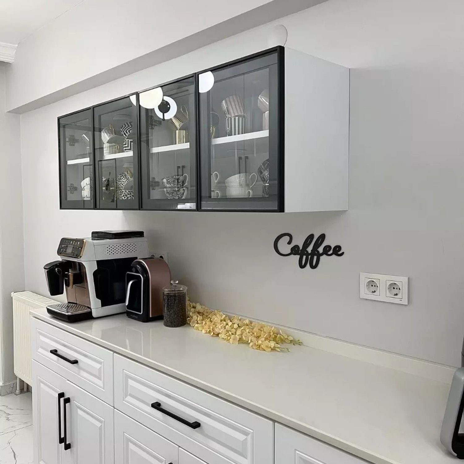 A modern kitchen coffee station with a sleek design featuring a white cabinet with black countertops and glass windowed upper cabinets displaying glassware and mugs. A coffee maker and capsules on the countertop add functionality, and a decorative "Coffee" sign hangs on the wall, enhancing the area's dedicated purpose.