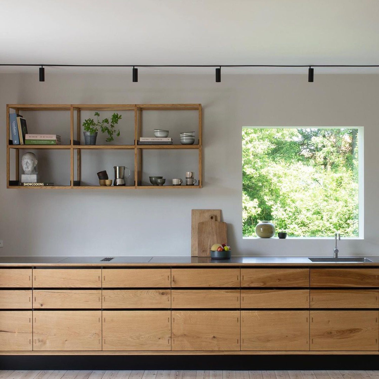 A sleek and modern kitchen featuring **natural wood cabinetry**, a **minimalistic shelf** with plants and ceramics, and a **bright window** overlooking greenery