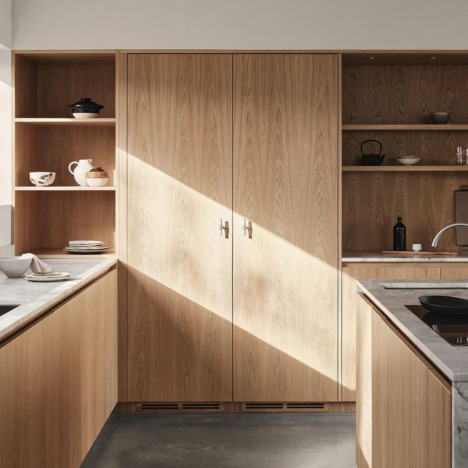 A modern kitchen showcasing light wood cabinets and open shelving, with sunlight casting a striking diagonal shadow across the surfaces