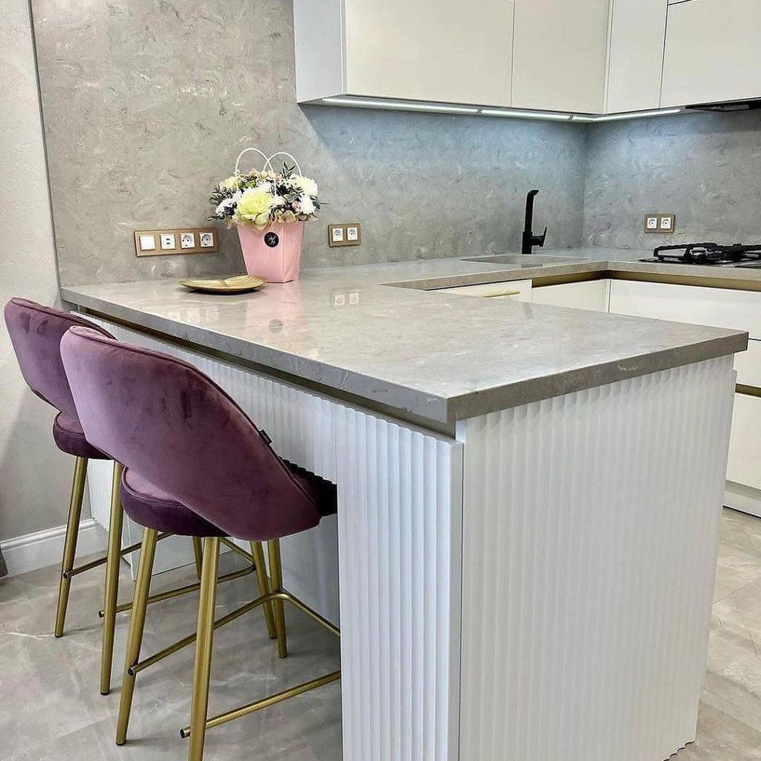 A modern kitchen featuring a sleek white island with a marble countertop, flanked by plush purple bar stools with golden legs.