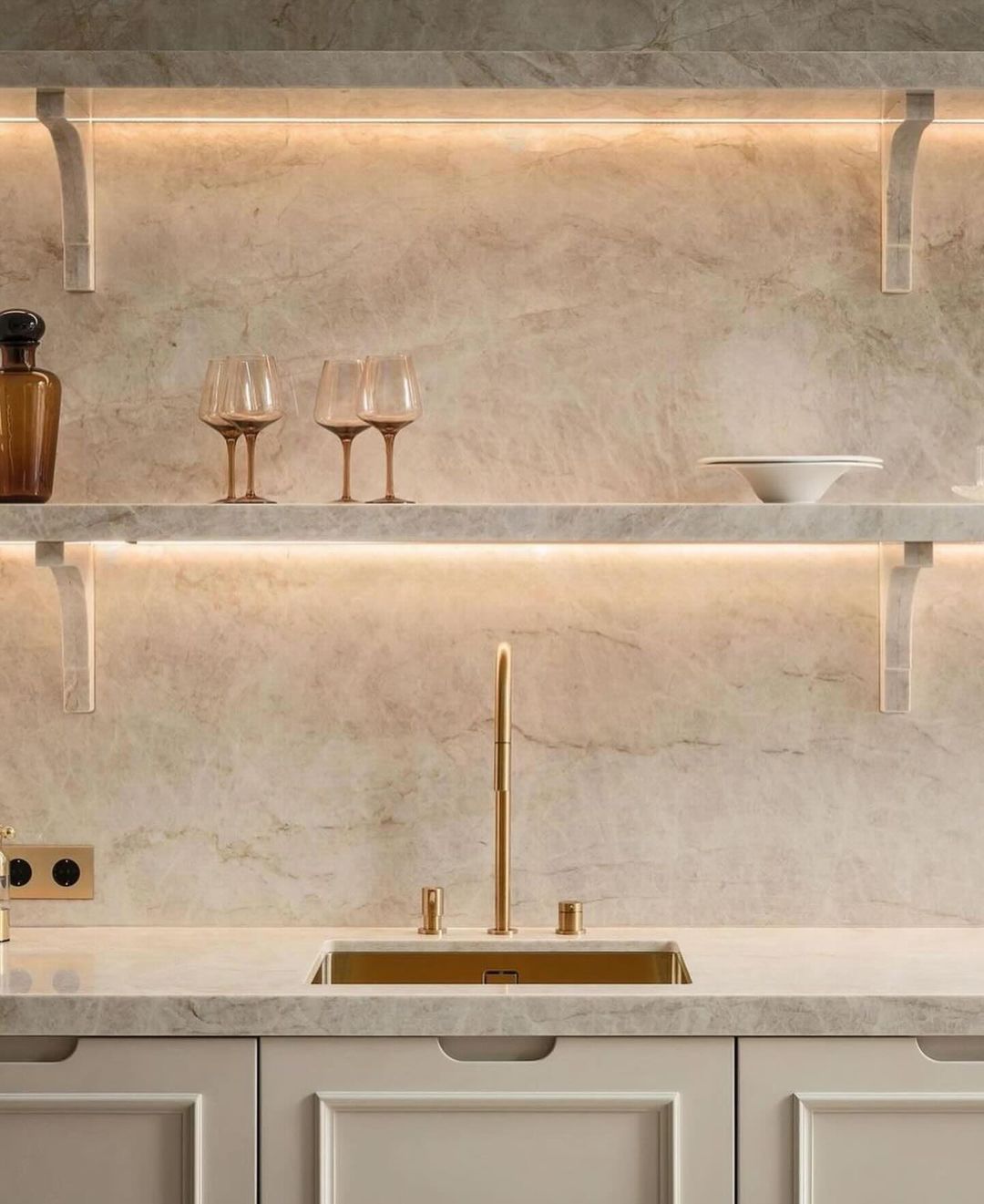 A tastefully designed kitchen featuring a marble backsplash and countertops with under-shelf lighting, accentuated by brass fixtures and stemware on display.