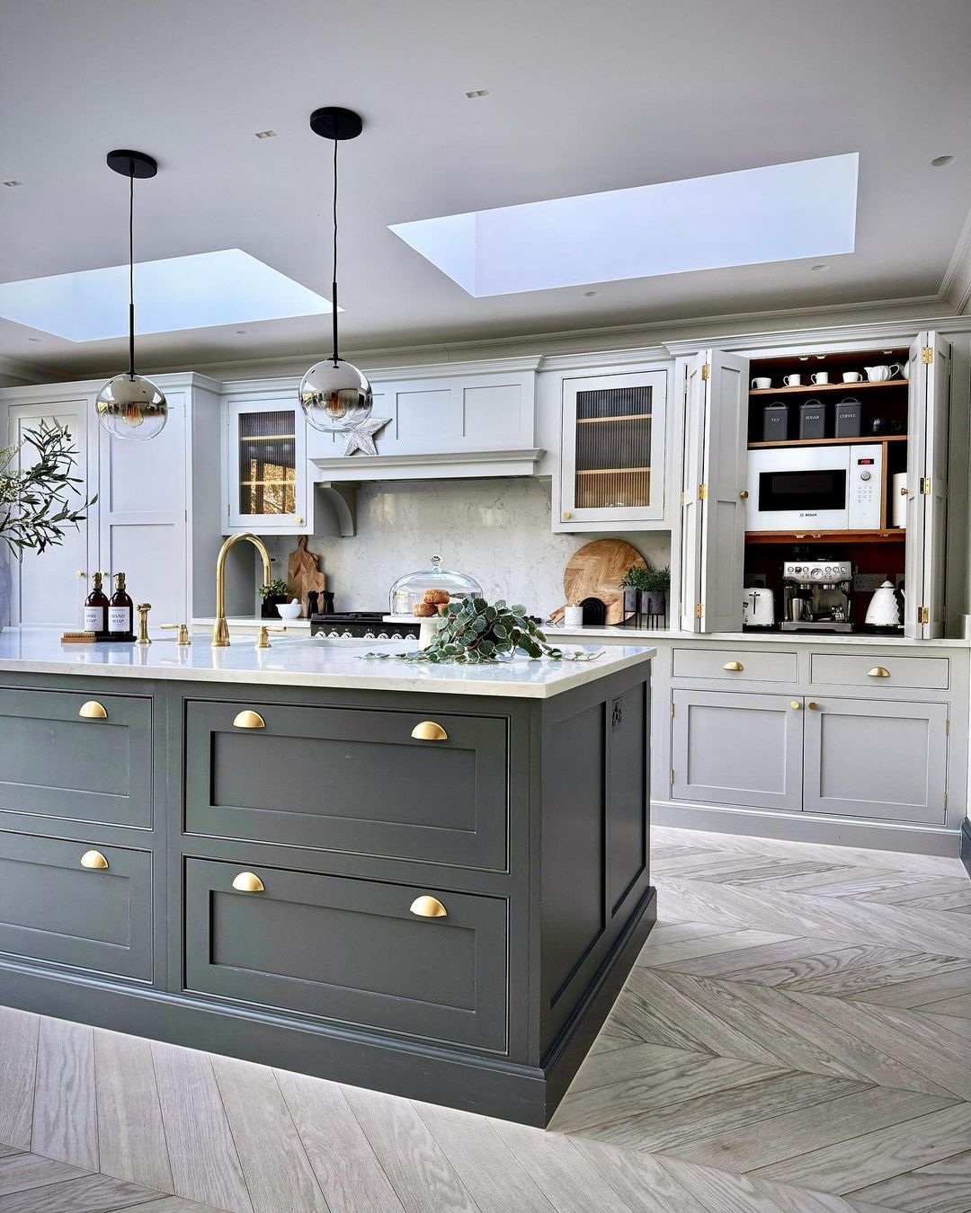 A sophisticated kitchen featuring a contrasting island in deep grey with brass handles against off-white cabinetry and marble accents.