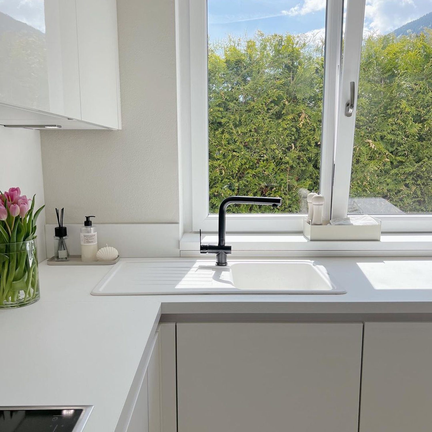 A modern kitchen space featuring an immaculate white theme with a sleek black faucet, set against a backdrop of lush greenery visible through the window.