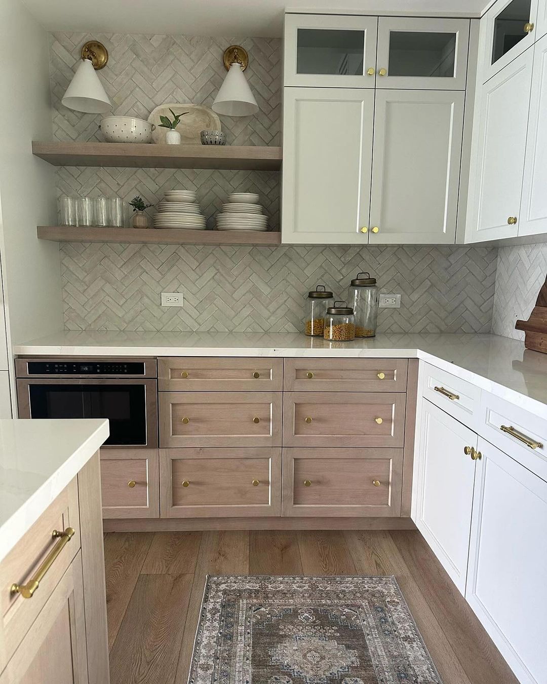 A modern kitchen featuring a blend of light wood drawers and white cabinetry, with gleaming gold hardware and herringbone backsplash