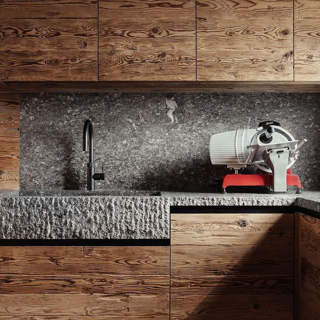 A modern kitchen featuring a combination of rustic wood cabinetry and textured dark stone surfaces, with a professional red and white meat slicer adding a pop of color to the neutral palette.