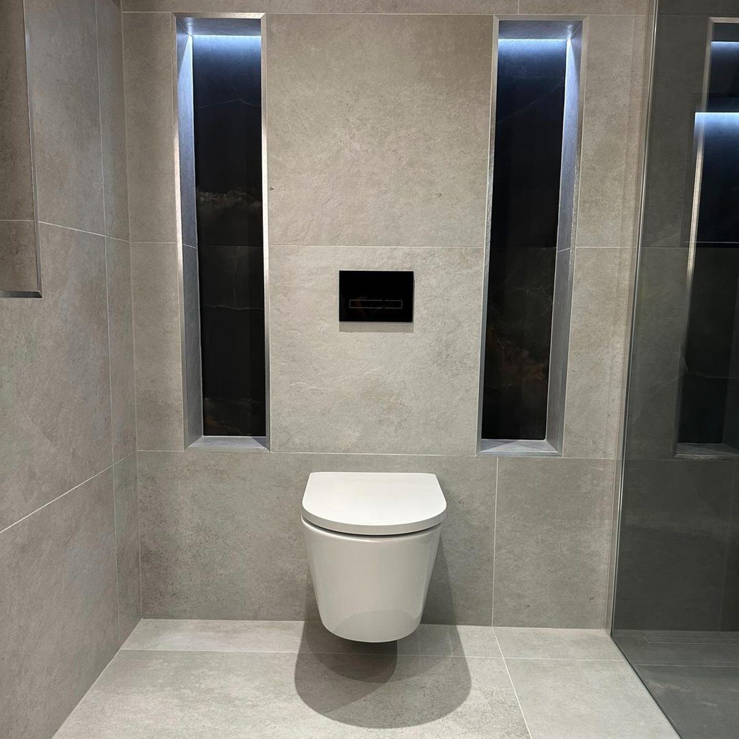 A modern minimalist bathroom featuring sleek grey tiles and an illuminated alcove above a wall-mounted toilet