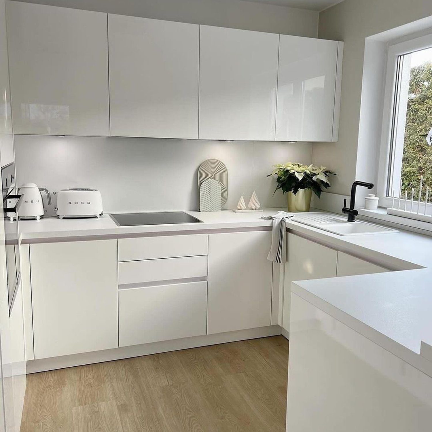 A modern, minimalist kitchen with sleek white cabinetry, featuring an undermount sink, matte black faucet, and a touch of greenery on the windowsill.