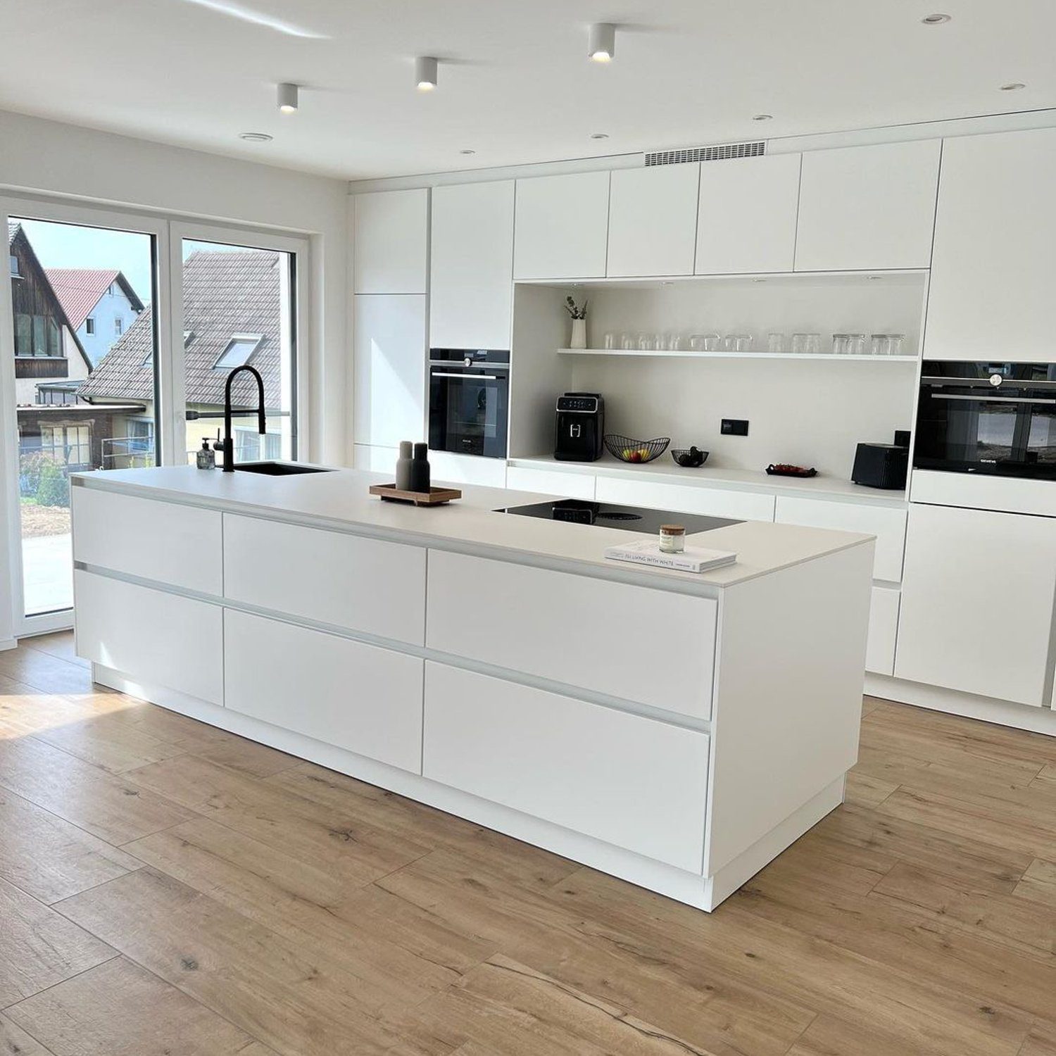 A modern kitchen featuring a minimalist design with sleek white cabinets, an island with a cooktop, and warm wooden floors